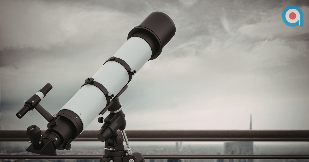 A white and black telescope on a tripod sits on a balcony, overlooking a hazy, distant cityscape under a cloudy sky