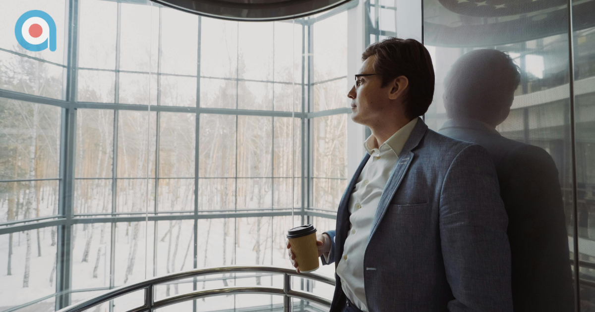 A man in a blazer looks out a large window at a snowy forest while holding a coffee cup.
