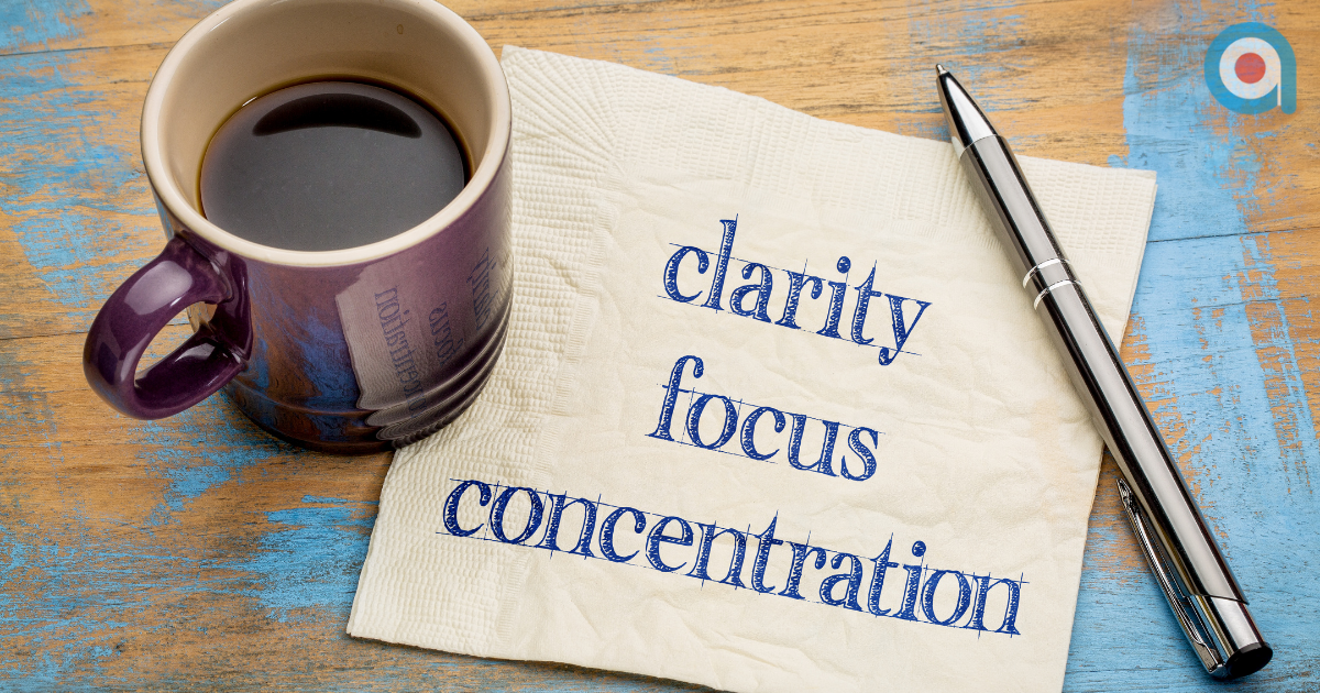 A cup of black coffee on a wooden table beside a pen and a napkin displaying the words clarity, focus and concentration.