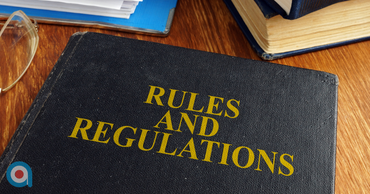 Close up of a black hardback book titled “Rules and Regulations” in yellow lettering, resting on a wooden desk with stacked books, papers and a pair of glasses nearby.